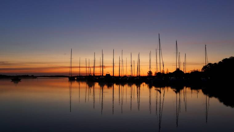 Aurinko värjää horisontin oranssiksi. Horisontin edessä kuvan keskellä on veneitä mustina. Tyyni vesi tumman sinistä. Kuva: Jarmo Vehkakoski.